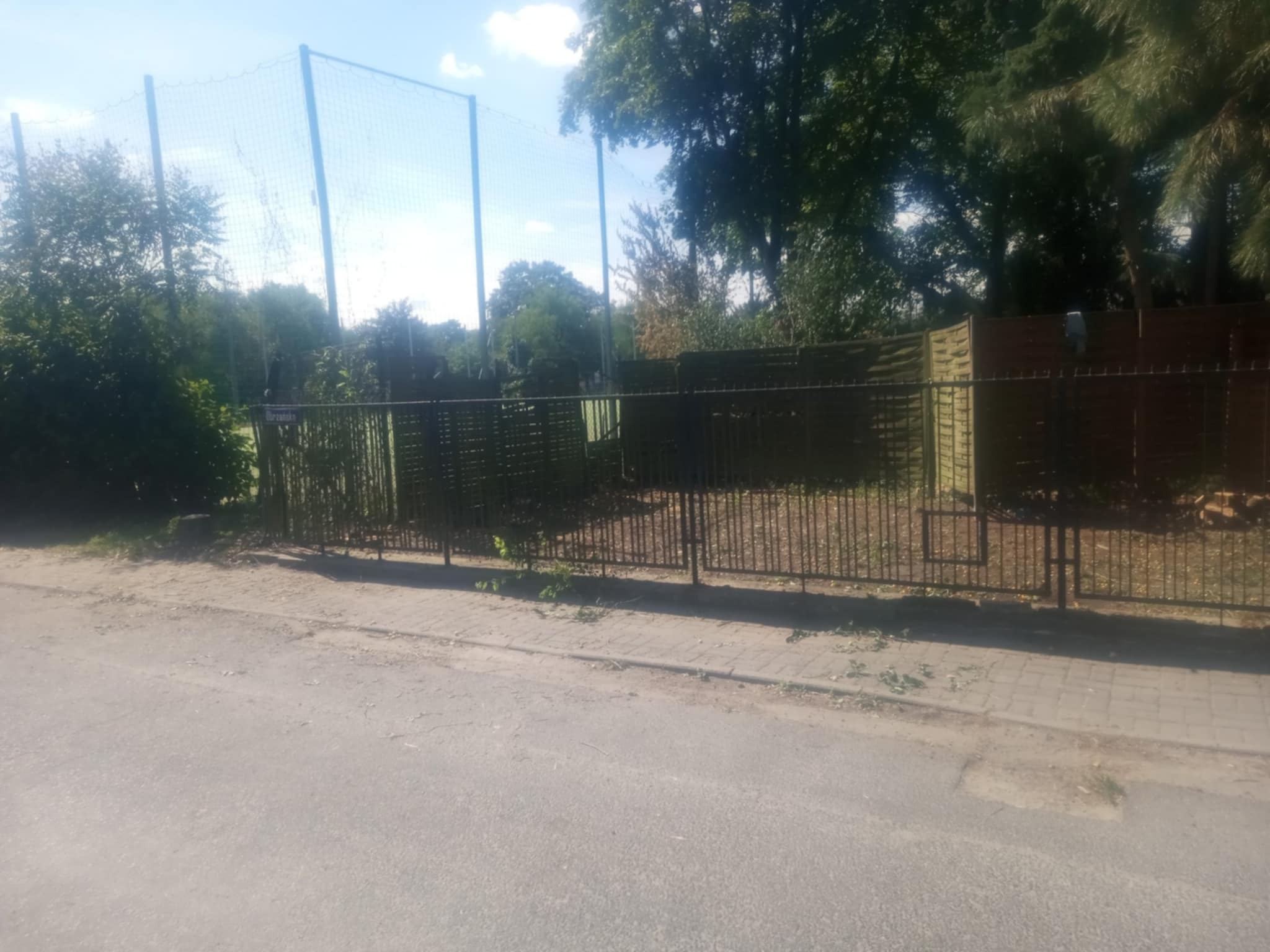 Metalowe ogrodzenie z bramą na tle zieleni i siatki, prawdopodobnie kort tenisowy. Świeżo przycięte gałęzie leżą na chodniku. Słoneczny dzień.