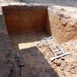 Firma Usługowa M-Bud Jakub Murzyn - Wykopany dół w ziemi, z widocznymi narzędziami (poziomica, młotek) i drabiną. U góry widać nogi osoby i statyw niwelacyjny, co sugeruje prace ziemne.