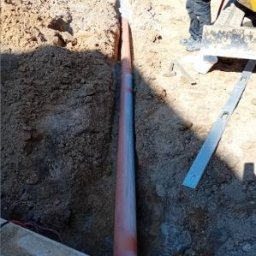 Firma Usługowa M-Bud Jakub Murzyn - Rów z pomarańczową rurą, częściowo zasypany ziemią. W tle mini koparka i poziomica. Widoczne przewody elektryczne. Słoneczny dzień.