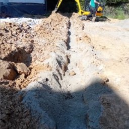 Firma Usługowa M-Bud Jakub Murzyn - Żółta mini-koparka wykopuje wąski rów w ziemi obok budynku w trakcie słonecznego dnia. Widoczny operator koparki i fragment rury w wykopie.