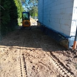 Firma Usługowa M-Bud Jakub Murzyn - Żółta mini koparka na placu budowy przy ścianie z bloczków. Widoczne ślady opon na ziemi i fragment zieleni w tle. Przygotowanie terenu pod budowę.