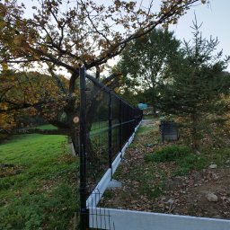 Firma Usługowa M-Bud Jakub Murzyn - Czarny, metalowy płot panelowy z podmurówką, widziany z perspektywy ziemi, otoczony jesiennym krajobrazem z drzewami i opadłymi liśćmi, w tle zielony trawnik.