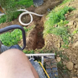 Wykop wąskiego rowu w ziemi z widoczną rurą drenażową i fragmentem minikoparki z perspektywy operatora, otoczenie trawa i zieleń.
