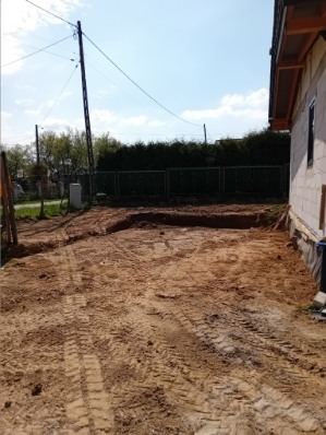 Wykop pod fundamenty domu jednorodzinnego, widoczne ślady opon maszyn budowlanych na piaszczystym terenie, budynek w trakcie budowy w tle.