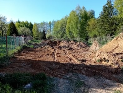 Przygotowany teren pod budowę, widoczne ślady opon, ziemia usypana w kopce, w tle drzewa i ogrodzenie. Słoneczny dzień.