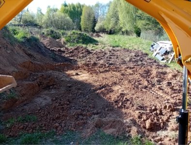 Żółta koparka w trakcie niwelacji terenu. Widok z kabiny na wyrównany teren z pozostałościami ziemi i skarpą. W tle drzewa i zielona trawa.