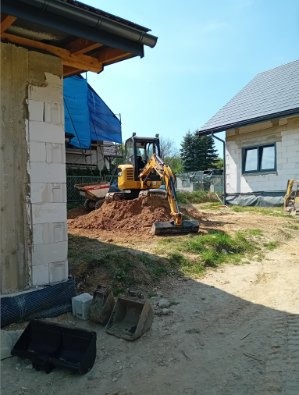 Żółta minikoparka na placu budowy, z wykopaną ziemią i widocznymi elementami konstrukcji domu w tle. W pierwszym planie leżą narzędzia.