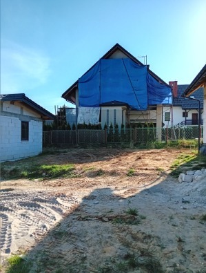 Przygotowany teren pod budowę domu, z widocznym fundamentem i zabezpieczonym dachem niebieską plandeką. Ziemia wyrównana, zarys działki wytyczony.
