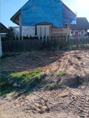 Dom w trakcie remontu, zabezpieczony niebieską plandeką, widoczne rusztowania i świeżo wyrównany teren z śladami opon, ogrodzenie panelowe.