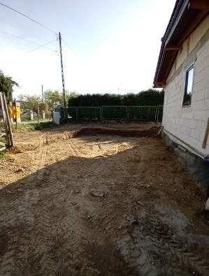 Przygotowany wykop pod fundamenty domu jednorodzinnego, widoczne ślady opon od ciężkiego sprzętu, w tle ogrodzenie i zieleń. Budynek w stanie surowym.