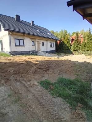 Nierówny teren przed domem jednorodzinnym z widocznymi śladami opon, ziemia usypana w kopce, dom w stanie surowym zamkniętym, błękitne niebo w tle.