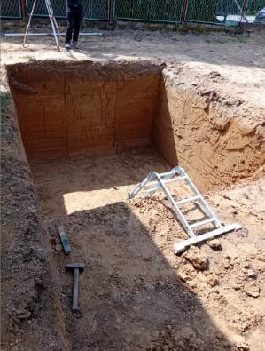 Wykopany dół w ziemi, z widocznymi narzędziami (poziomica, młotek) i drabiną. U góry widać nogi osoby i statyw niwelacyjny, co sugeruje prace ziemne.