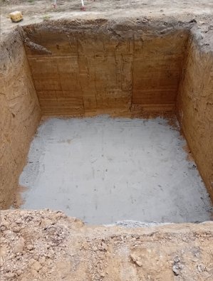 Kwadratowy wykop w ziemi z wyrównanym dnem. Ściany wykopu warstwowe, widoczne ślady po narzędziach. Dół wykopu wypełniony jasnoszarą substancją.