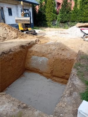 Wykop pod fundamenty z widoczną żółtą mini koparką i przyczepą z piaskiem w tle, na działce obok domu w trakcie budowy. Dół ma równe, wyprofilowane ściany.
