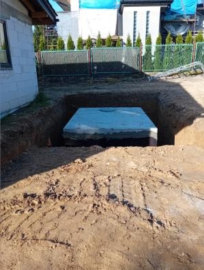 Betonowy zbiornik w wykopie na posesji, otoczony ziemią, z widocznym ogrodzeniem i budynkiem w tle. Etap prac ziemnych przed zasypaniem.