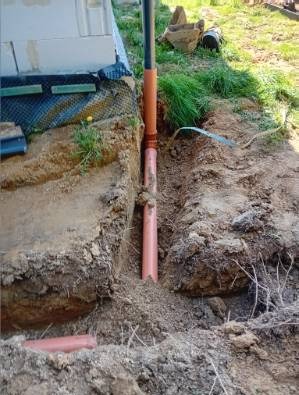 Pomarańczowa rura kanalizacyjna w wykopie, widoczne elementy izolacji budynku i ziemia. Rura pionowo wkopana w ziemię, na dnie wykopu leży kolejna rura.