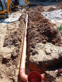 Wykonany drenaż: rów z ułożonymi rurami drenarskimi z tworzywa sztucznego, widoczny żółty minikoparka w tle, grunt piaszczysty.