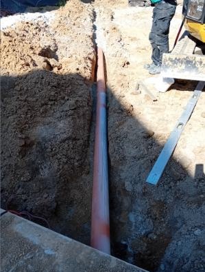 Rów z pomarańczową rurą, częściowo zasypany ziemią. W tle mini koparka i poziomica. Widoczne przewody elektryczne. Słoneczny dzień.