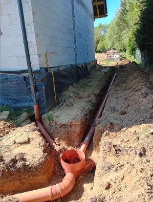 Instalacja drenażowa z pomarańczowych rur w wykopie przy fundamencie budynku, widok z góry, czarna folia izolacyjna na fundamencie.