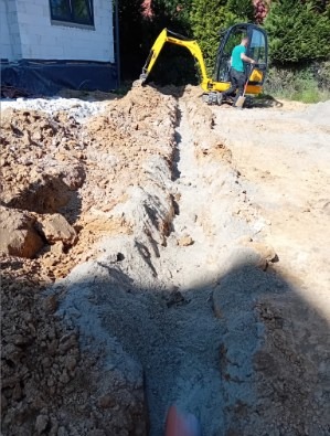 Żółta mini-koparka wykopuje wąski rów w ziemi obok budynku w trakcie słonecznego dnia. Widoczny operator koparki i fragment rury w wykopie.