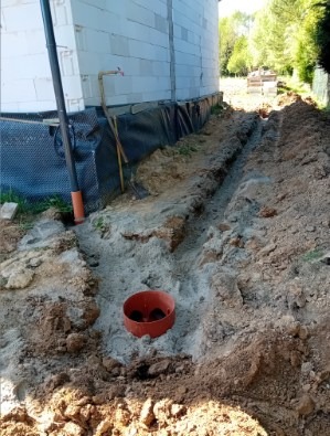Wykopany rów wzdłuż fundamentów budynku z widoczną rurą drenażową. Fundament zabezpieczony folią hydroizolacyjną. Prace ziemne w trakcie realizacji.