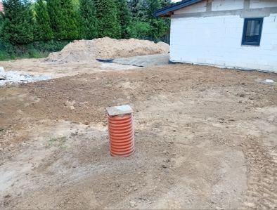 Teren po niwelacji z widocznym pomarańczowym pionowym elementem drenażowym. W tle sterta ziemi i budynek w trakcie budowy. Ślady opon na ziemi.