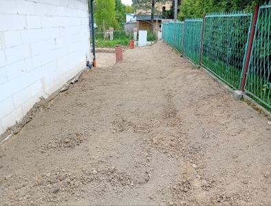 Przygotowany teren pod budowę, wyrównana ziemia wzdłuż białej ściany budynku i zielonego ogrodzenia. Widoczne ślady po pracach ziemnych.
