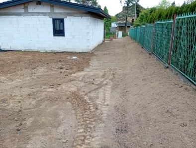 Przygotowany teren pod budowę lub utwardzenie, widoczne ślady opon, budynek w tle i zielone ogrodzenie po prawej stronie kadru.