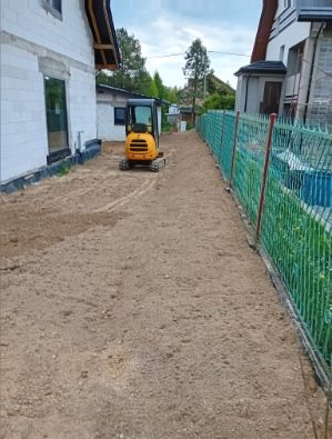 Żółta mini koparka na wyrównanym terenie obok zielonego ogrodzenia i domu w trakcie prac ziemnych. Ślady opon widoczne na piasku.