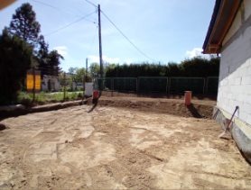 Przygotowany teren pod budowę z widocznymi słupkami i częściowo ogrodzeniem w tle, w słoneczny dzień. Widoczny fragment budynku z oknem.