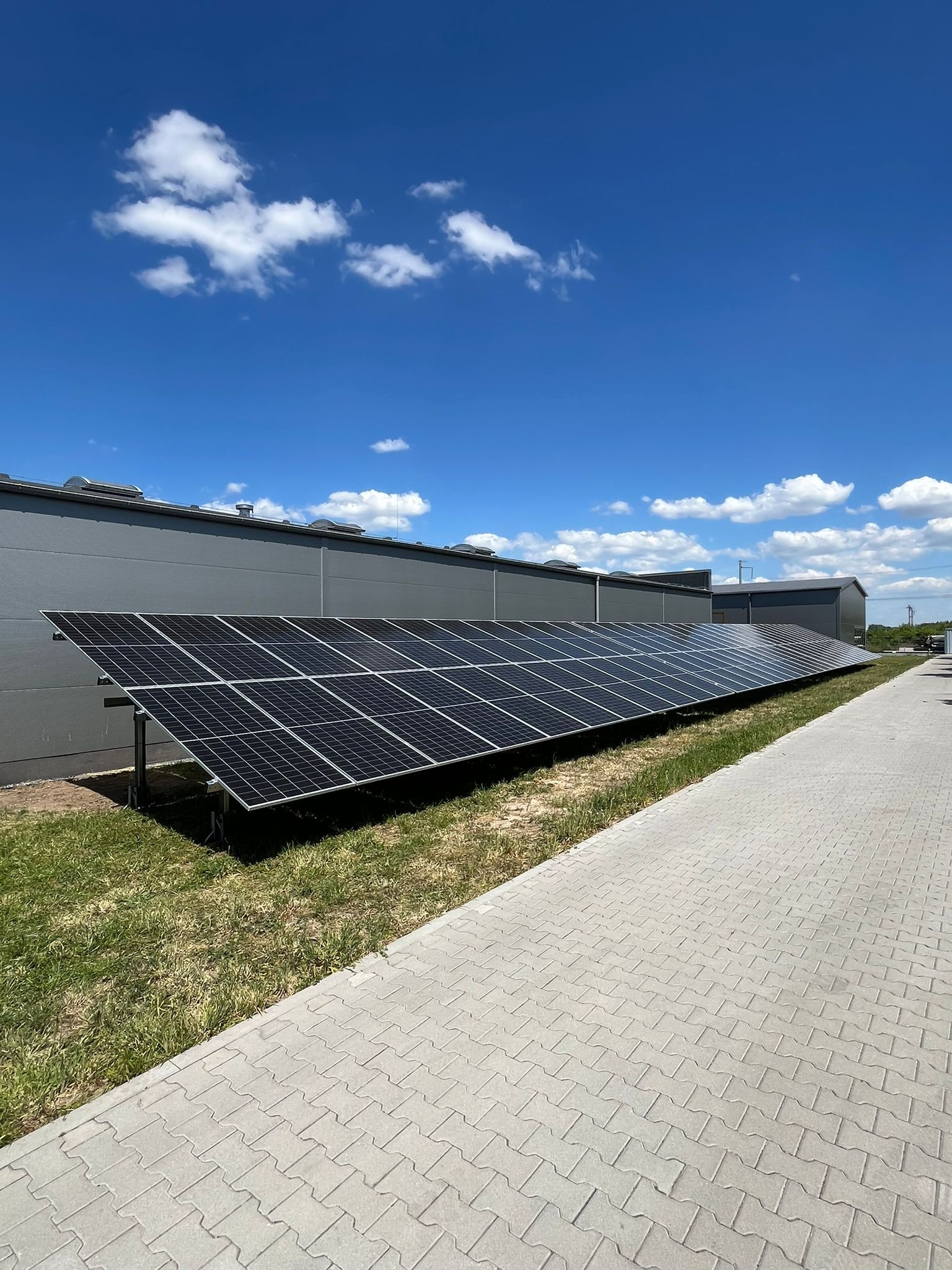 Instalacja fotowoltaiczna przy budynku przemysłowym w słoneczny dzień. Panele ustawione w rzędzie na trawie, obok chodnik z kostki brukowej. Błękitne niebo z chmurami.