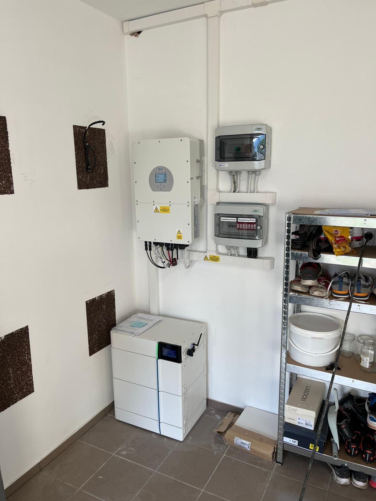 Instalacja magazynu energii w garażu: biały modułowy akumulator, falownik i rozdzielnice na ścianie, metalowy regał z butami obok.