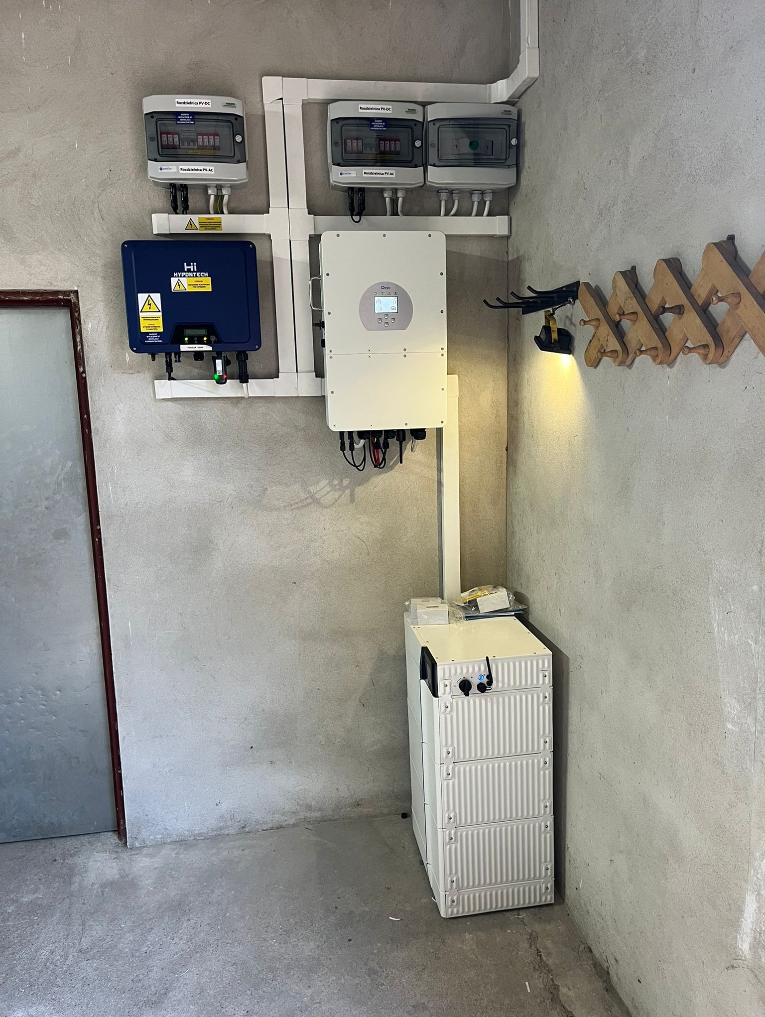 Instalacja fotowoltaiczna w pomieszczeniu: biały inwerter Deye, niebieski Hypontech, rozdzielnice PV-DC/AC i modułowy magazyn energii na tle surowej ściany.