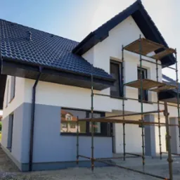 Dom jednorodzinny z rusztowaniem, pomalowany na biało i szaro, z czarnym dachem. Świeżo pomalowana elewacja, widoczne detale wykończeniowe. Słoneczny dzień.