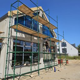 FLOT Docieplenia, Elewacje, Wykończenia - Budowa domu w Wierzchucinie: elewacja w trakcie tynkowania, widoczne rusztowania, okna zabezpieczone folią. Prace na wysokości.