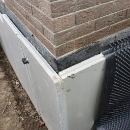 FLOT Docieplenia, Elewacje, Wykończenia - Fragment elewacji budynku w trakcie prac: białe płyty izolacyjne i folia kubełkowa przy fundamencie, ceglany narożnik. Widoczne detale mocowania.