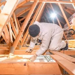 FLOT Docieplenia, Elewacje, Wykończenia - Pracownik w kombinezonie ochronnym ociepla poddasze wełną mineralną, widoczne drewniane elementy konstrukcji dachu i rolki izolacji w tle.