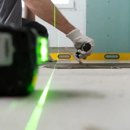 FLOT Docieplenia, Elewacje, Wykończenia - Układanie płytek na podłodze z użyciem poziomicy laserowej. Widoczna ręka w rękawicy rozprowadzająca klej. W tle poziomica tradycyjna i płyta kartonowo-gipsowa.