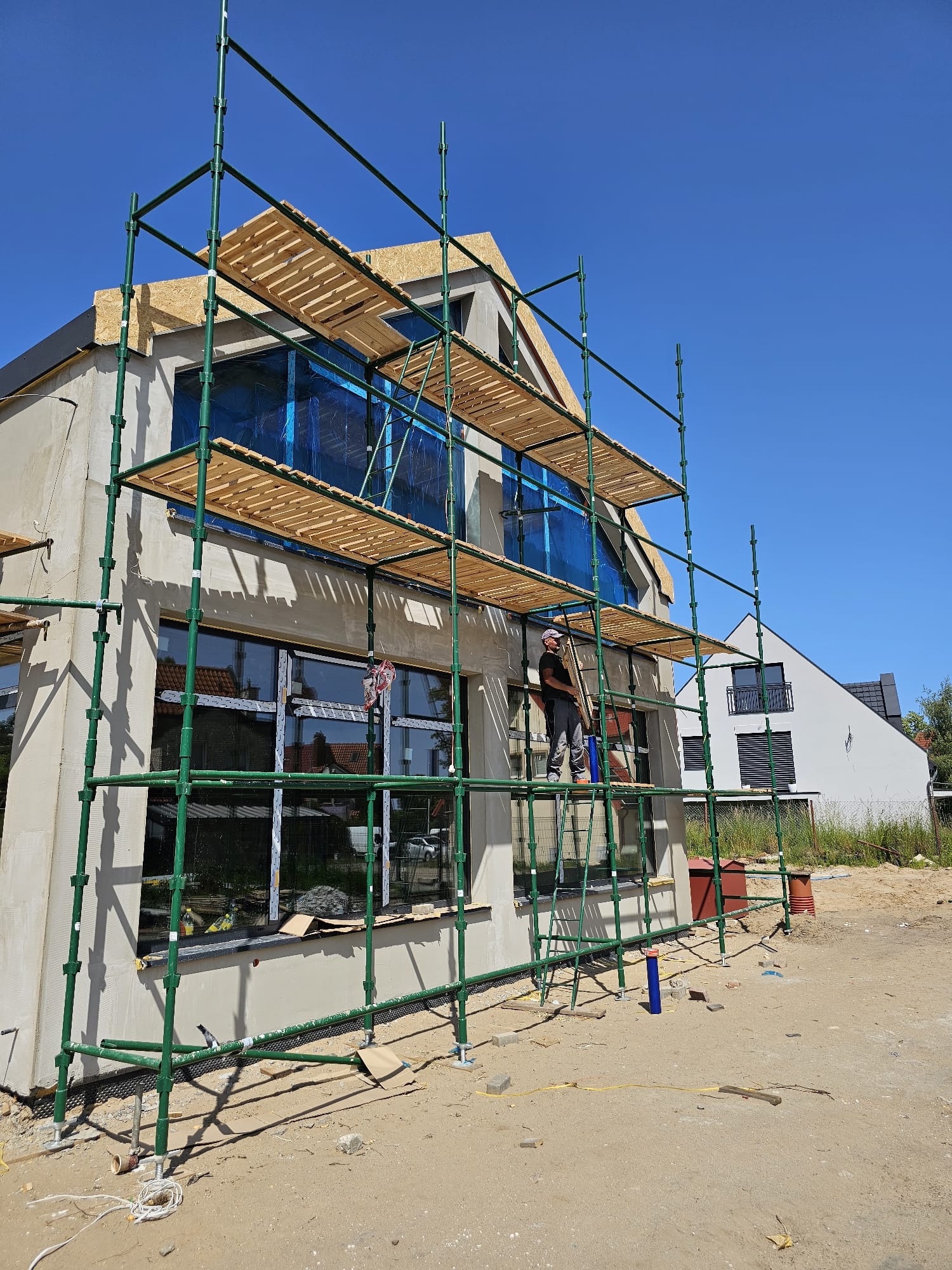 Budowa domu w Wierzchucinie: elewacja w trakcie tynkowania, widoczne rusztowania, okna zabezpieczone folią. Prace na wysokości.