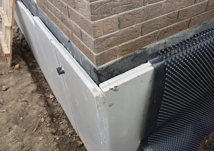 Fragment elewacji budynku w trakcie prac: białe płyty izolacyjne i folia kubełkowa przy fundamencie, ceglany narożnik. Widoczne detale mocowania.