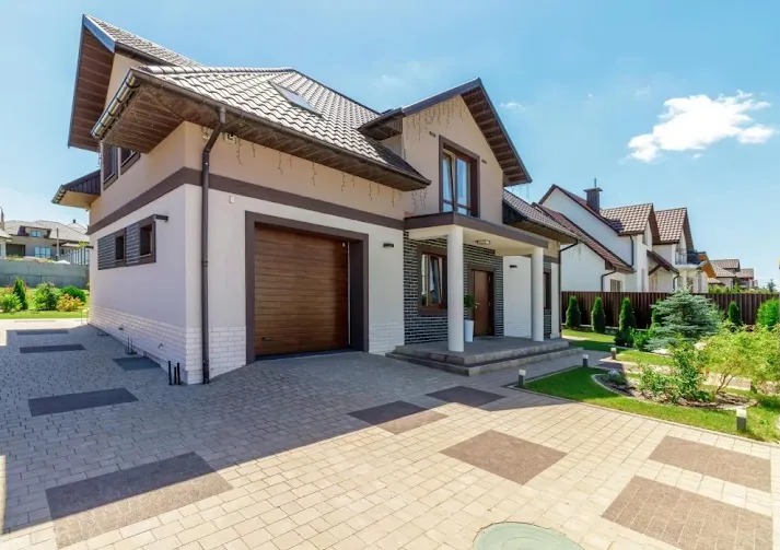 Dwukondygnacyjny dom z garażem i podjazdem z kostki, beżowa elewacja z ciemnobrązowymi akcentami, brązowa brama garażowa, białe kolumny przy wejściu.