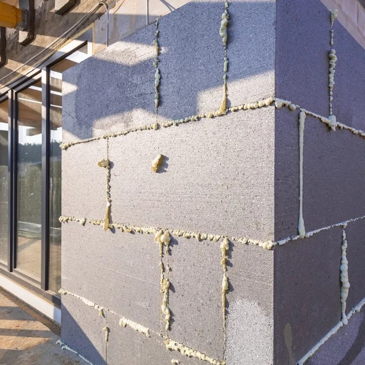 Szara ściana z bloczków z widoczną pianką montażową, narożnik budynku w surowym stanie, okno w tle, budowa domu w trakcie realizacji.