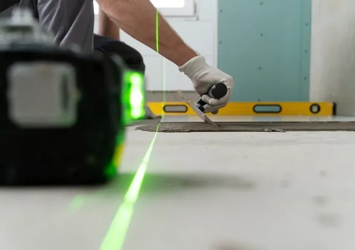 Układanie płytek na podłodze z użyciem poziomicy laserowej. Widoczna ręka w rękawicy rozprowadzająca klej. W tle poziomica tradycyjna i płyta kartonowo-gipsowa.