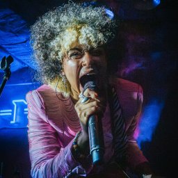 mKanikphoto - Energetyczna wokalistka z kręconymi włosami i blond pasemkami, w różowej bluzce, śpiewa do mikrofonu na scenie, z gitarą i niebieskim neonem w tle.