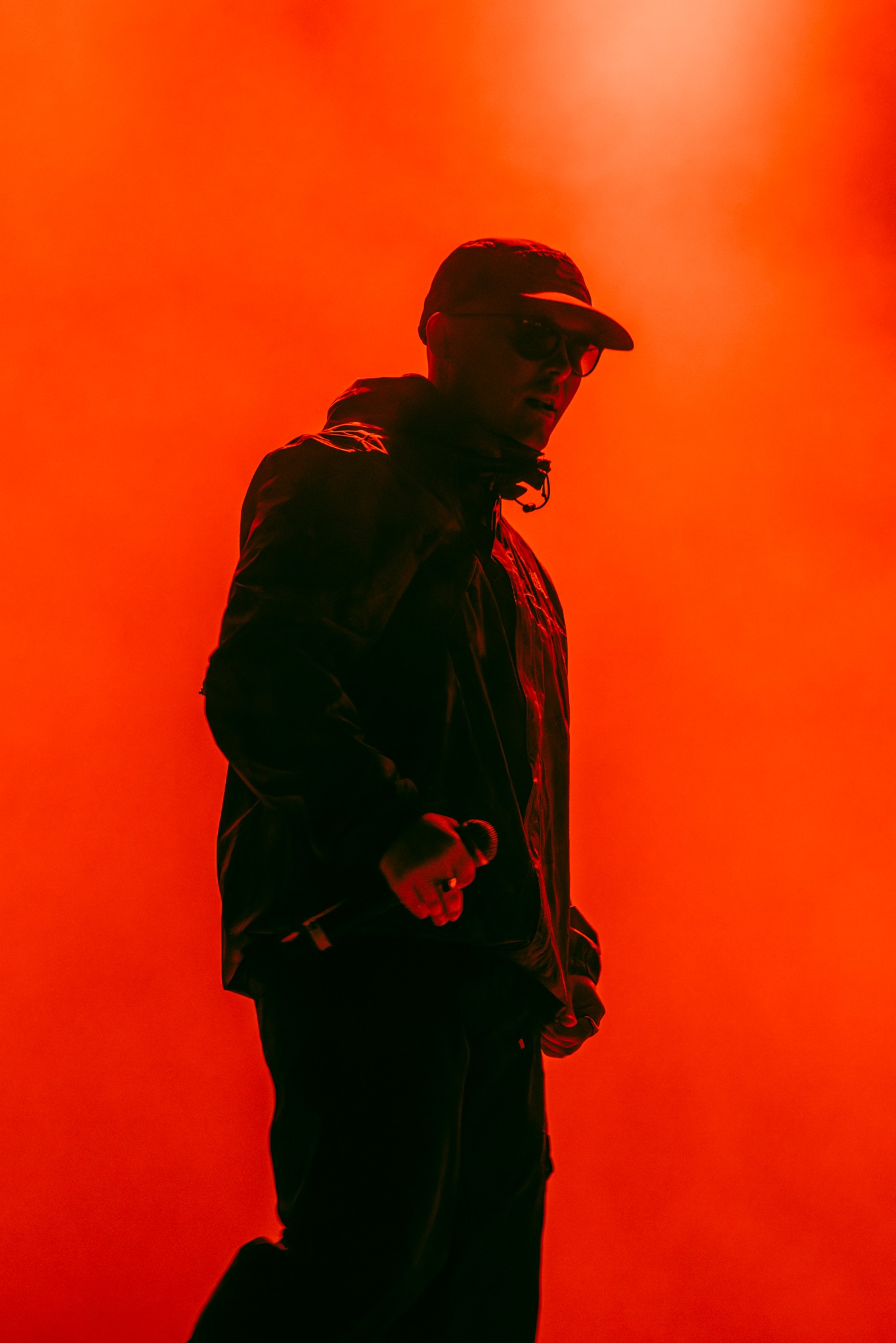 Silueta mężczyzny w czapce i okularach, trzymającego mikrofon na tle intensywnego, czerwonego oświetlenia scenicznego. Kontrastowe światło tworzy dramatyczny efekt.