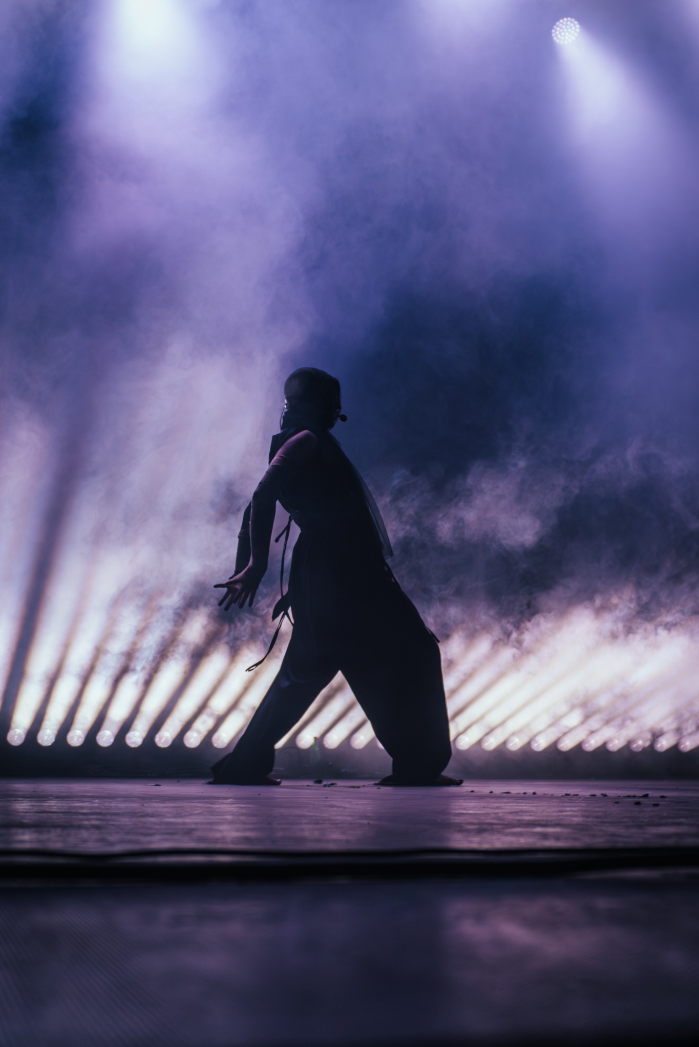 Sylwetka tancerki w ruchu, oświetlona fioletowym światłem reflektorów i otoczona dymem na scenie. Dynamiczne ujęcie z mikrofonem nagłownym.