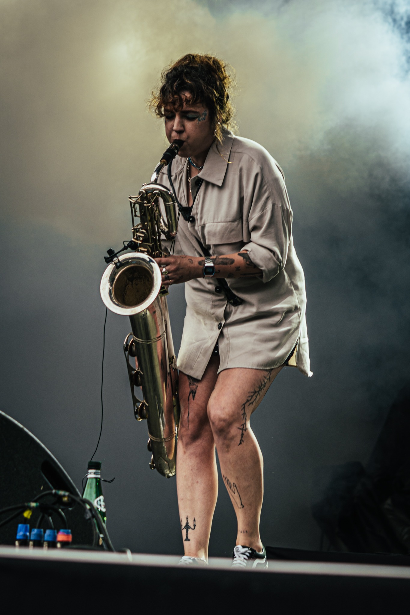 Kobieta z tatuażami gra na saksofonie na scenie w oparach dymu, ubrana w beżową koszulę, z widocznymi butami sportowymi i butelką wody w tle.