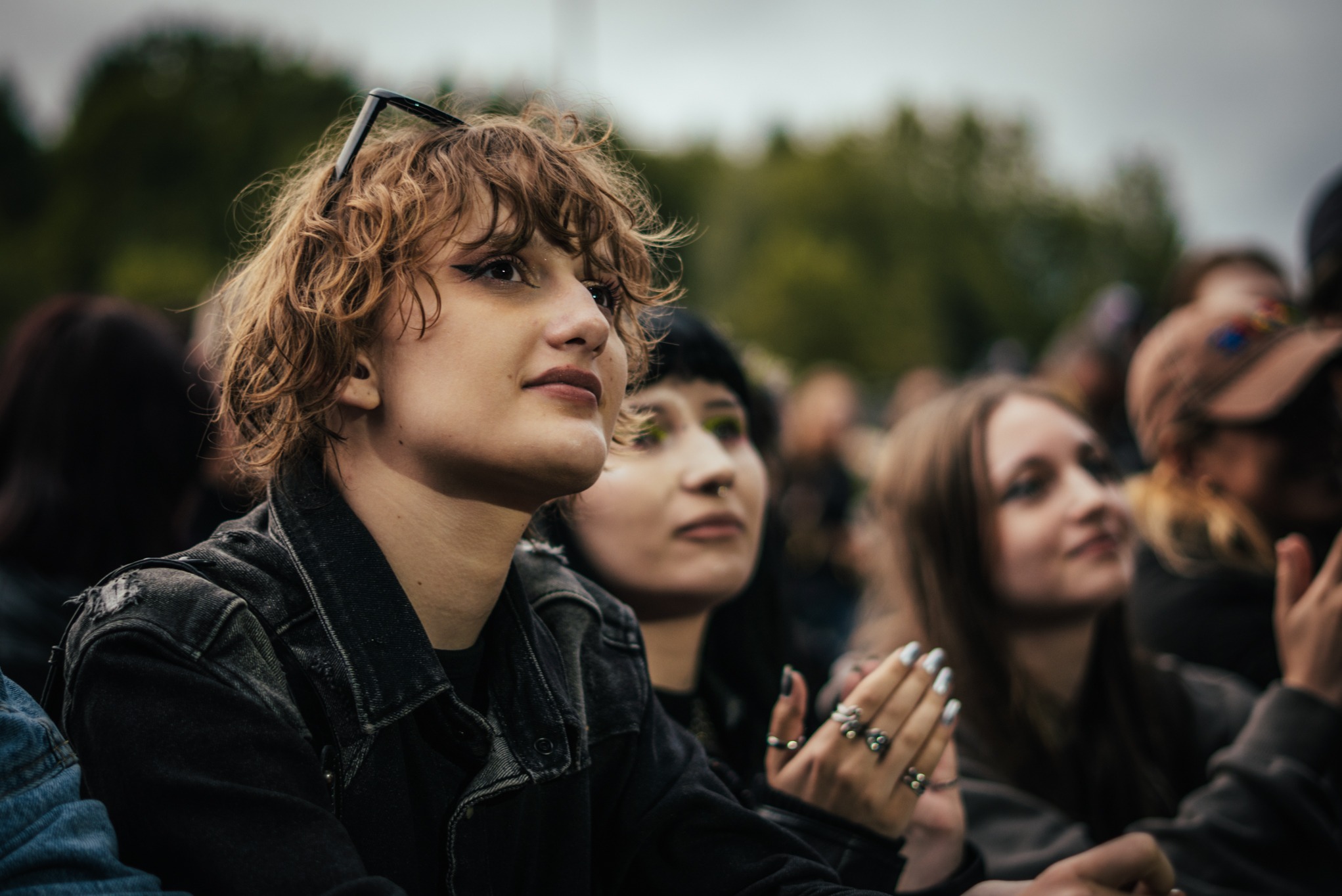 Tłum młodych ludzi na koncercie; w pierwszym planie dziewczyna z kręconymi włosami, w czarnej kurtce dżinsowej, z okularami na głowie, patrzy w górę.