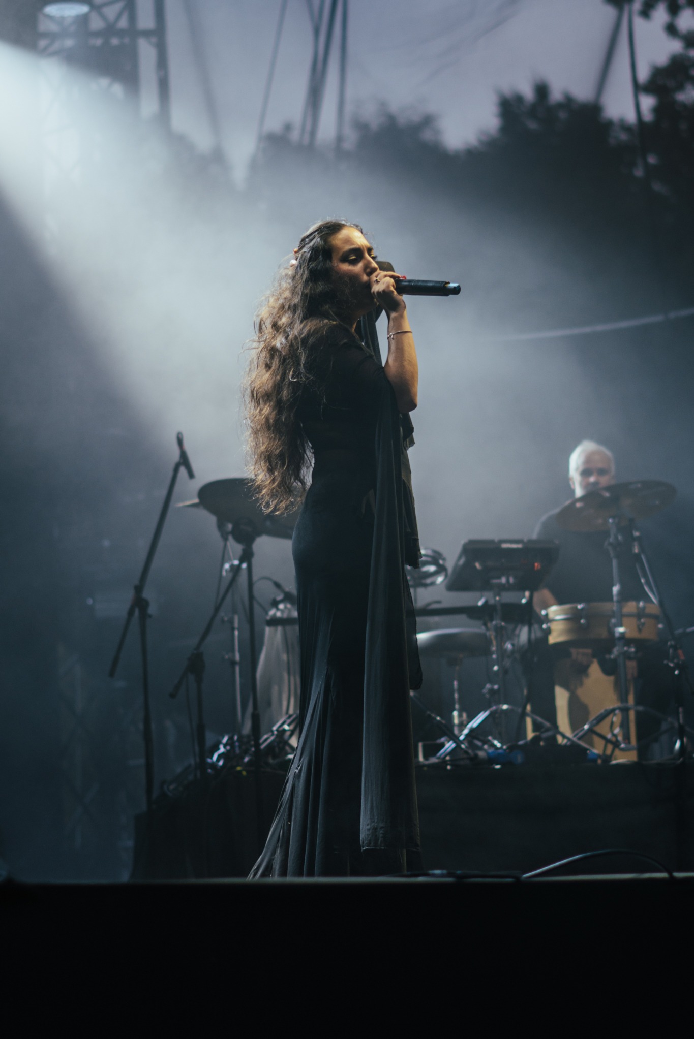 Wokalistka w długiej czarnej sukni śpiewa do mikrofonu na scenie, w tle perkusista i sztuczne dymy, oświetlenie sceniczne. Ujęcie z dołu, ciemna tonacja.