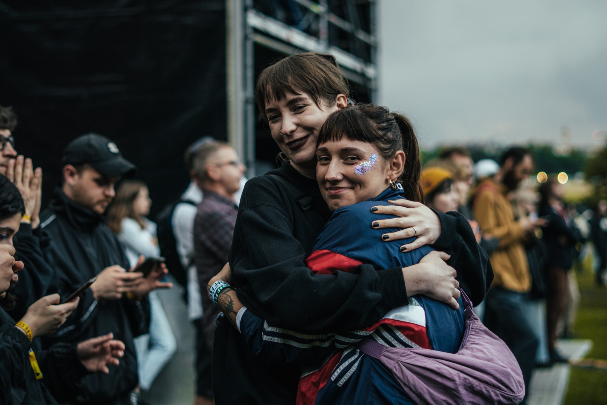 Dwie kobiety obejmują się na tle tłumu podczas wydarzenia plenerowego, jedna z brokatem na policzku, druga z ciemną szminką i kolczykiem w nosie.
